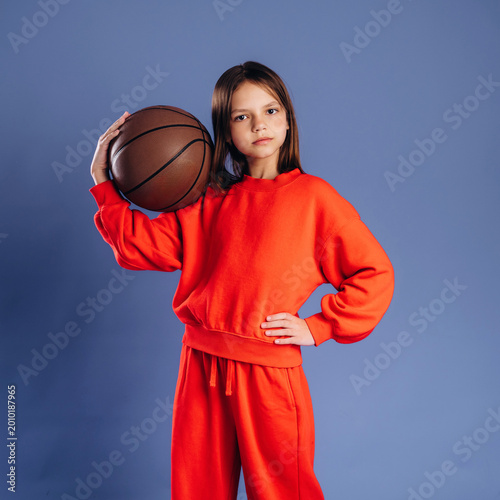 Young girl wearing bright red sportswear holds a basketball in one hand while standing confidently against a blue background