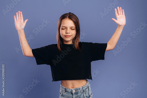 Young girl with brown hair wearing a black crop top and denim shorts raises her arms in a relaxed pose against a solid blue background