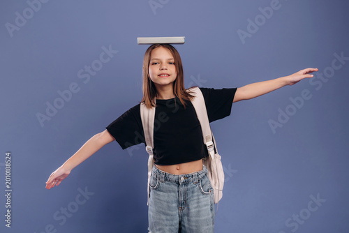 Young girl balancing a book on her head while standing with arms outstretched, wearing a black t-shirt and denim jeans against a solid blue background