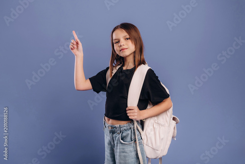 Young girl with long brown hair wearing a black shirt and blue jeans holds a white backpack and points upwards against a solid blue background