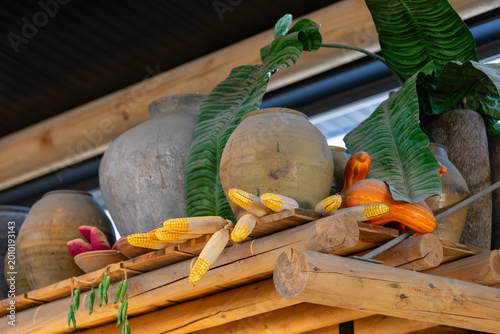 Rustic decorative display with artificial corn, clay pots, wooden beams and green tropical leaves. Warm interior detail with natural materials and harvest-inspired styling.