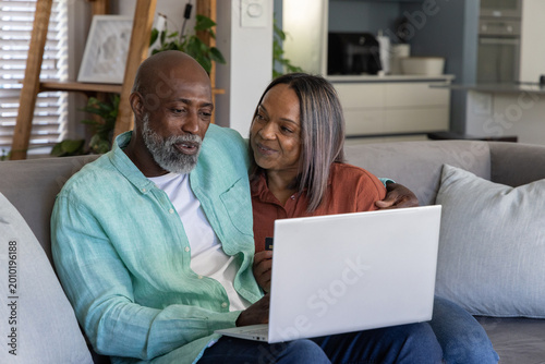 Couple sitting together on gray sofa reviewing online bills on white laptop, woman holding card
