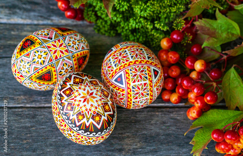 Three hand-painted Ukrainian Easter eggs (pysanky) featuring intricate geometric patterns and solar symbols. The eggs are arranged on a rustic wooden surface next to red viburnum (kalyna) berries 