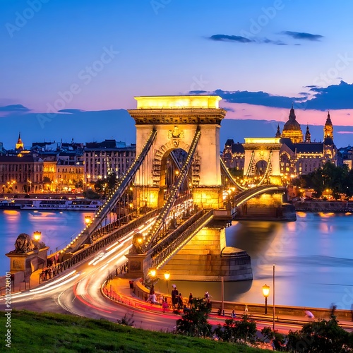 Illuminated suspension bridge with flowing traffic lights over a wide river at twilight, featuring city buildings