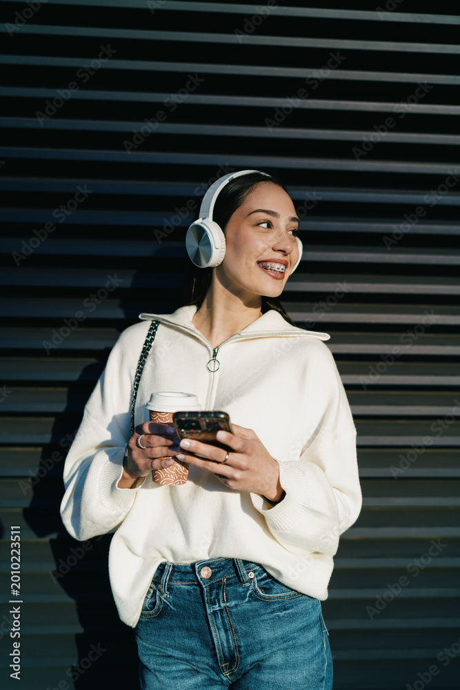 Fototapeta premium Cheerful young woman with braces using mobile phone in headphones and holding takeaway coffee. Girl enjoying digital connectivity, streaming music or chatting in contemporary urban setting.