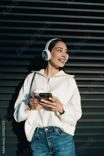Cheerful young woman with braces using mobile phone in headphones and holding takeaway coffee. Girl enjoying digital connectivity, streaming music or chatting in contemporary urban setting.