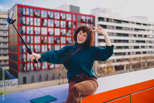 Valokuva Young woman pouts and flashes peace sign while taking a selfie using a smartphone on a selfie stick, embracing playful digital self-portraits in modern mobile communication culture