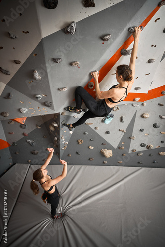 Woman supports climbing partner during bouldering session indoors. Photo for safety, encouragement, teamwork, strength, gym membership marketing, sport communication
