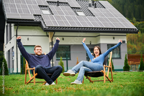 Couple relaxing on deck chairs near house with solar panels, raising hands and celebrating eco friendly lifestyle outdoors. Concept of renewable energy, freedom and sustainable living
