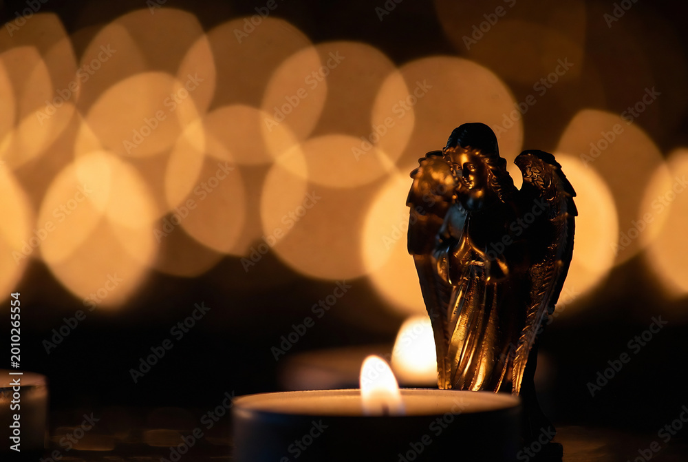 Fototapeta premium A bronze angel statue rests on a surface with blurred candlelight, creating a warm and serene atmosphere.
