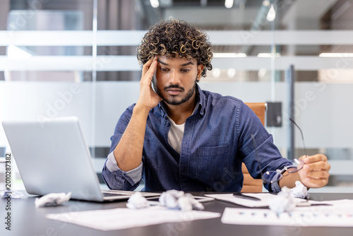 Картината върху платно Thoughtful young Indian man sitting at a desk in the office, holding his head in