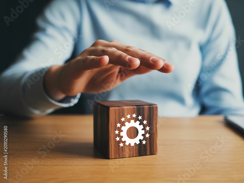 Businessman protecting wooden block with gear icon on desk, corporate security concept