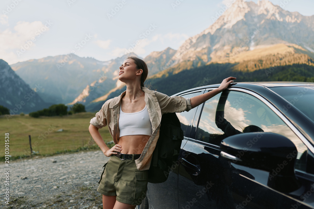 Fototapeta premium Young woman in casual outfit leans on car outdoors enjoys fresh air with mountains in background expresses freedom and lifestyle in nature travel concept.