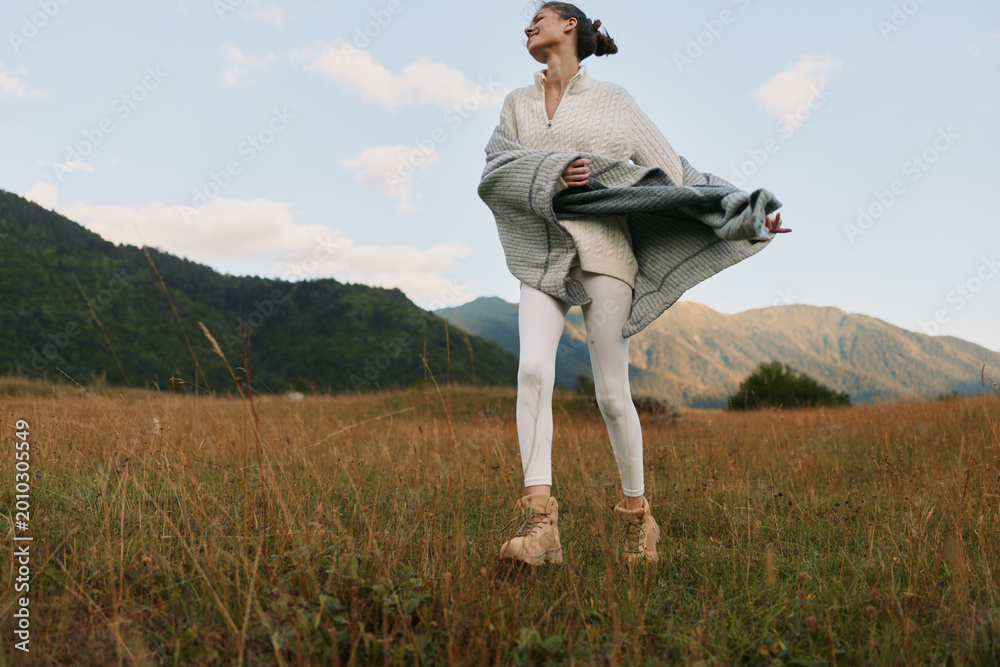 Naklejka premium Woman in a light sweater and coat stands in a grassy field near mountains, enjoying fresh air and outdoor lifestyle amid nature.
