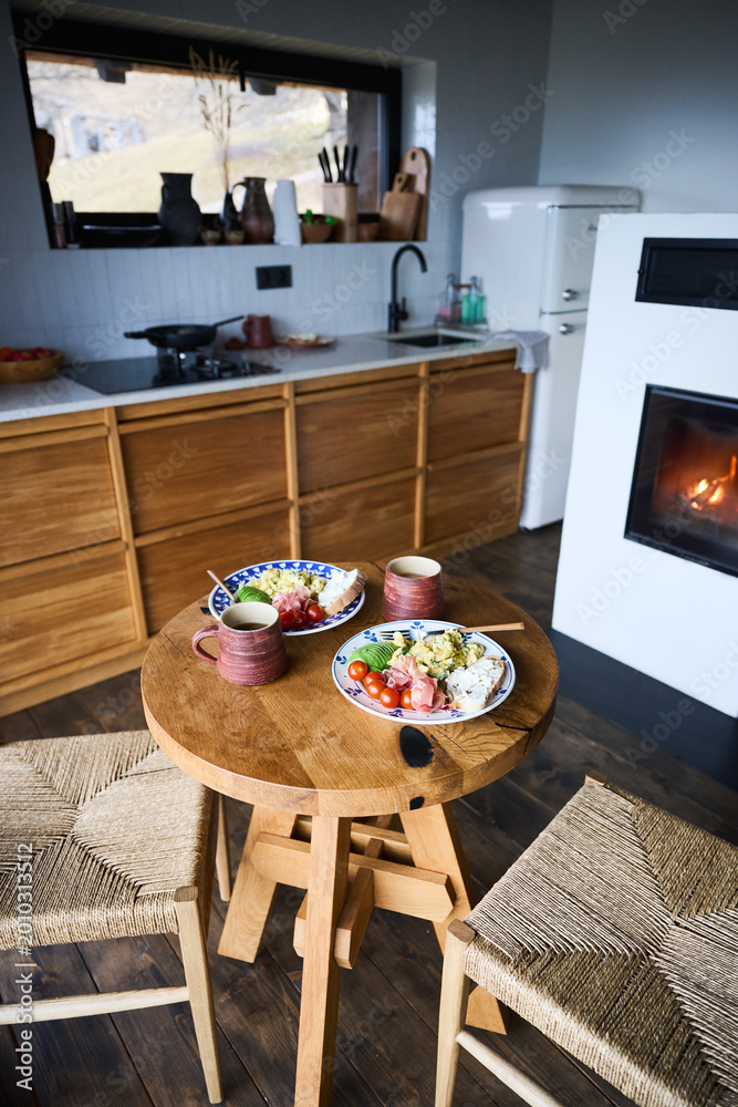 Naklejka premium Healthy breakfast setup in cozy modern kitchen with fireplace, wooden table set for two with eggs, avocado, prosciutto and coffee, concept of home lifestyle, comfort, slow living