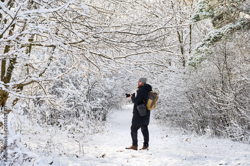 Fototapeta premium Winter photographer stands on snowy forest path with camera, surrounded by frosted branches and quiet woodland. Useful for travel marketing, winter tourism, outdoor advertising, and lifestyle concepts