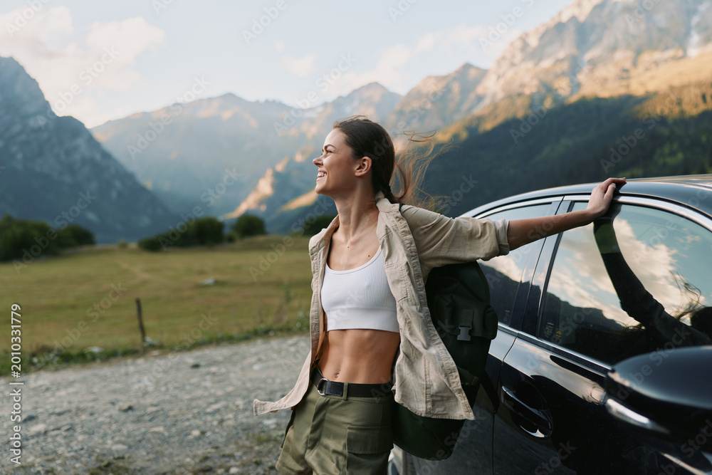Naklejka premium A woman stands beside a car in a wide meadow, arms outstretched, savoring freedom and nature as mountains rise behind, capturing a carefree, outdoors lifestyle moment
