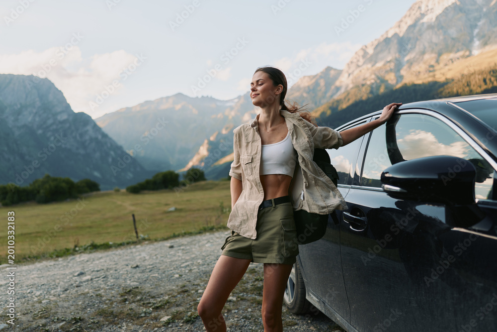 Fototapeta premium A woman leans against a dark car in a mountain valley, wearing a cropped top and casual jacket, embodying travel, freedom, and outdoor lifestyle in a bright landscape.