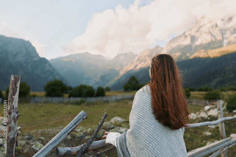 Naklejka premium Woman with long hair in a knit sweater stands beside a wooden fence gazing at distant mountains, serene outdoor lifestyle scene, nature and wide open space.