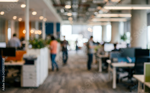 A modern office interior with blurred figures engaged in work. Desks are arranged along an open layout with plants and natural light.