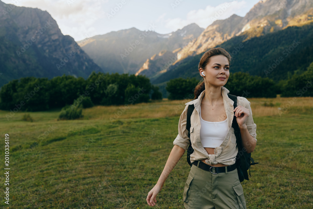 Naklejka premium Cheerful woman outdoors, walking through a sunlit meadow with mountains in the distance, casual hiking gear, backpack, relaxed expression, natural colors, fresh breeze, serene mood