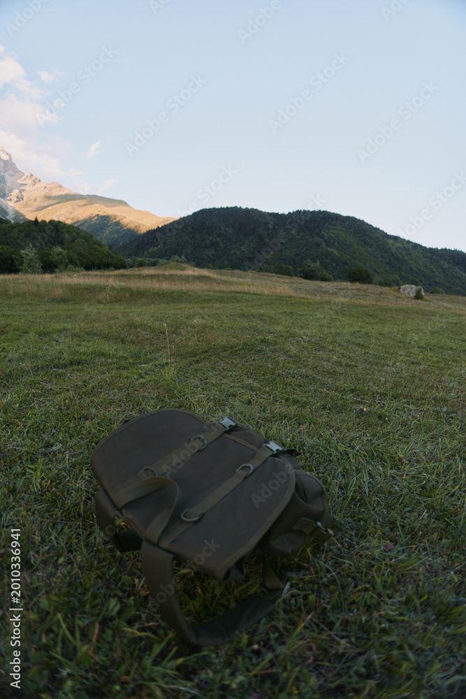 Naklejka premium A lone backpack lies on a grassy meadow with distant hills and a clear sky, conveying outdoor travel, exploration, and a sense of nature escape.