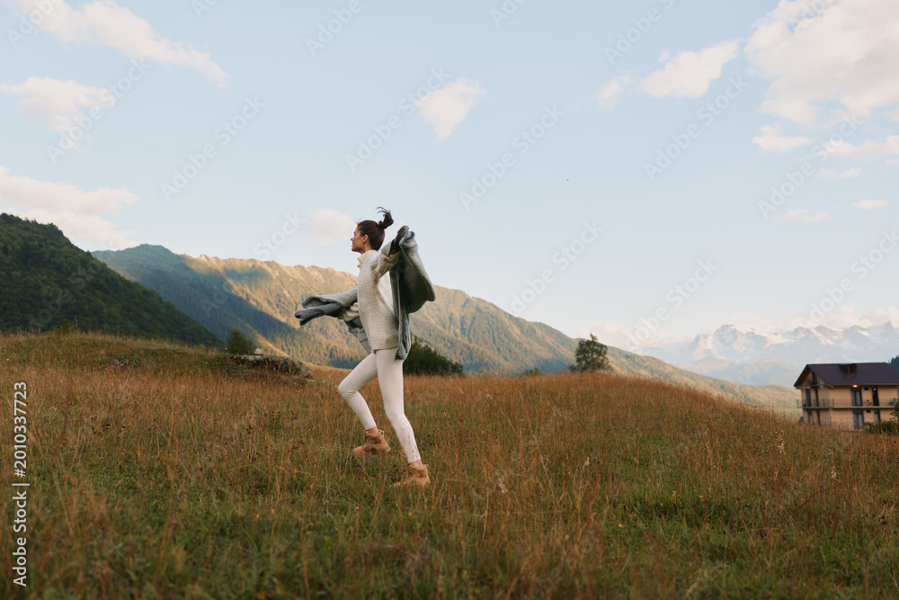 Naklejka premium Young woman with backpack hikes across a grassy field toward distant mountains, radiating carefree adventure and energy in an open landscape at golden hour with soft breeze.