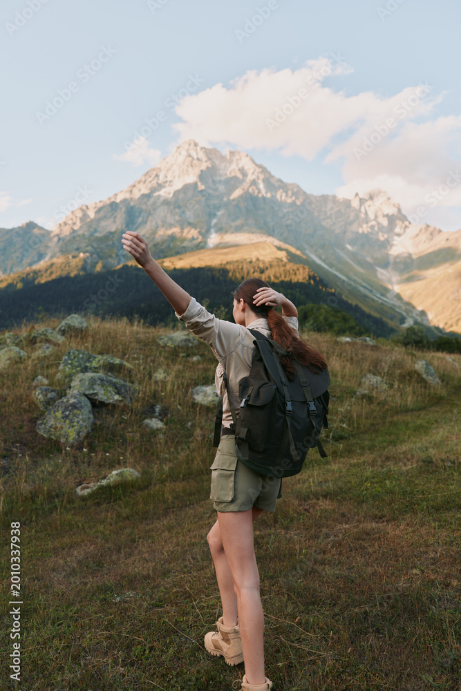 Naklejka premium A woman with backpack hikes through alpine meadow, arms raised, rugged mountains in the background under a bright sky, conveying adventure and outdoor lifestyle.