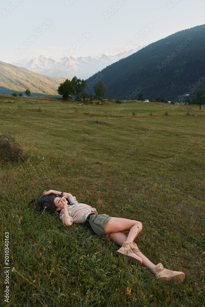 Naklejka premium Relaxed woman lying in a green meadow with distant hills, enjoying nature and sunshine. Casual outfit highlights outdoor lifestyle and a peaceful, carefree moment and mood.