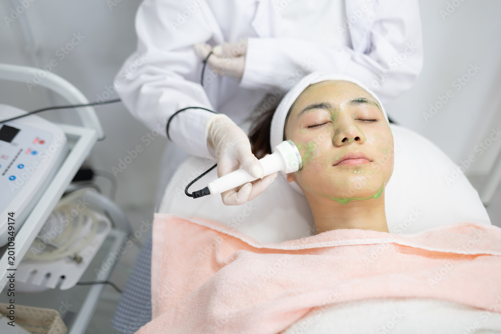 Naklejka premium Woman relaxes during facial treatment. Professional uses device gently on her face. Her eyes are closed, face calm and peaceful. Clinic environment appears clean and modern. Ideal for beauty, skincare