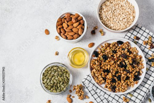 Homemade granola clusters and raw ingredients for cooking breakfast. A top view of plate with granola cereal, bowls of oats, nuts, seeds, honey on white table. Flat lay banner with copy space.