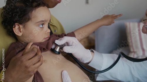 Medium close up handheld shot of male medical worker in gloves and white coat listening to breath of African American toddler with stethoscope, sitting with mother during home visit