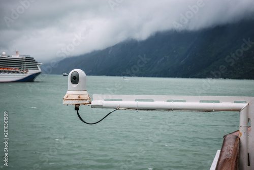 maritime thermal imaging camera mounted on a ship's railing with a cruise ship and misty Alaskan mountains in the background.