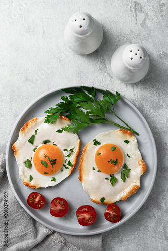 A home-cooked breakfast: two fried eggs garnished with chopped herbs, a bunch of fresh parsley, and cherry tomatoes, on a minimalist gray plate against a light textured background.