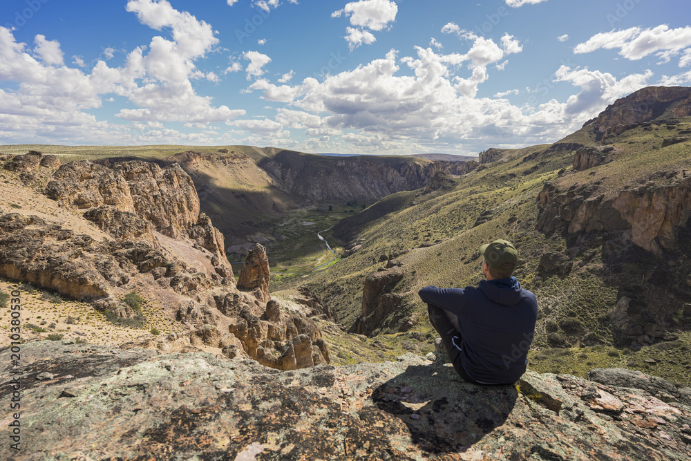 Fototapeta premium Wide angle view of man sitting on rock looking at epic valley of Pinturas River Canyon. Parque Patagonia, Argentina