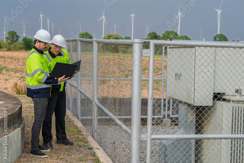 Two professional engineers using laptop for analysis at wind turbine farm power plant substation. Quality technical inspection and maintenance for sustainable renewable energy electricity grid.