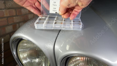 Closeup of a man’s hand taking an old type glass fuse from a box of varied current amp ratings, resting on the bodywork of a silver classic car in a garage workshop.