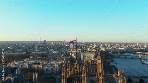 Waving Union Jack flag on top of Victoria Tower in London with Big Ben and River Thames while camera orbits the Houses of Parliament on a winter sunset