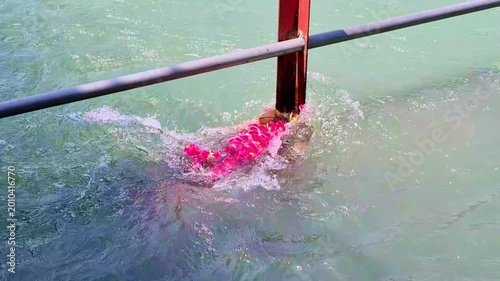 A vibrant garland of pink flowers and a small ritual lamp drift through the swirling, pale green waters of Ganges at Haridwar, momentarily catching against a metal safety railing near the riverbank.