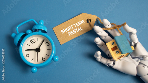 Robot hand holding a miniature house and a short term rental tag next to a blue alarm clock representing AI in real estate and booking management.