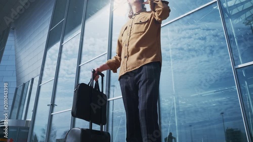 A curly-haired traveler pauses on the airport sidewalk, his luggage beside him, as he talks on his mobile phone. The glass terminal and soft daylight create a calm, busy travel backdrop.
