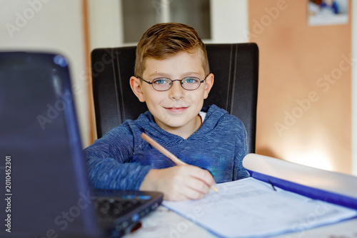 Happy healthy kid boy with glasses making school homework at home with notebook. Interested child writing essay with helping of internet. concetrated schoolchildren concept