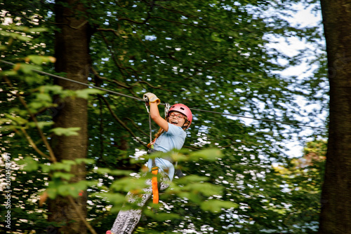Happy little girl climbing rope course in forest adventure park, child wearing helmet and harness enjoying outdoor activity, real life moment. School child having fun.