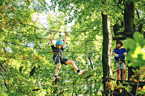 Two teenager boys climbing rope course in forest adventure park, teenagers wearing helmets and harnesses enjoying outdoor challenge, real life. Brothers and best friends outdoors.