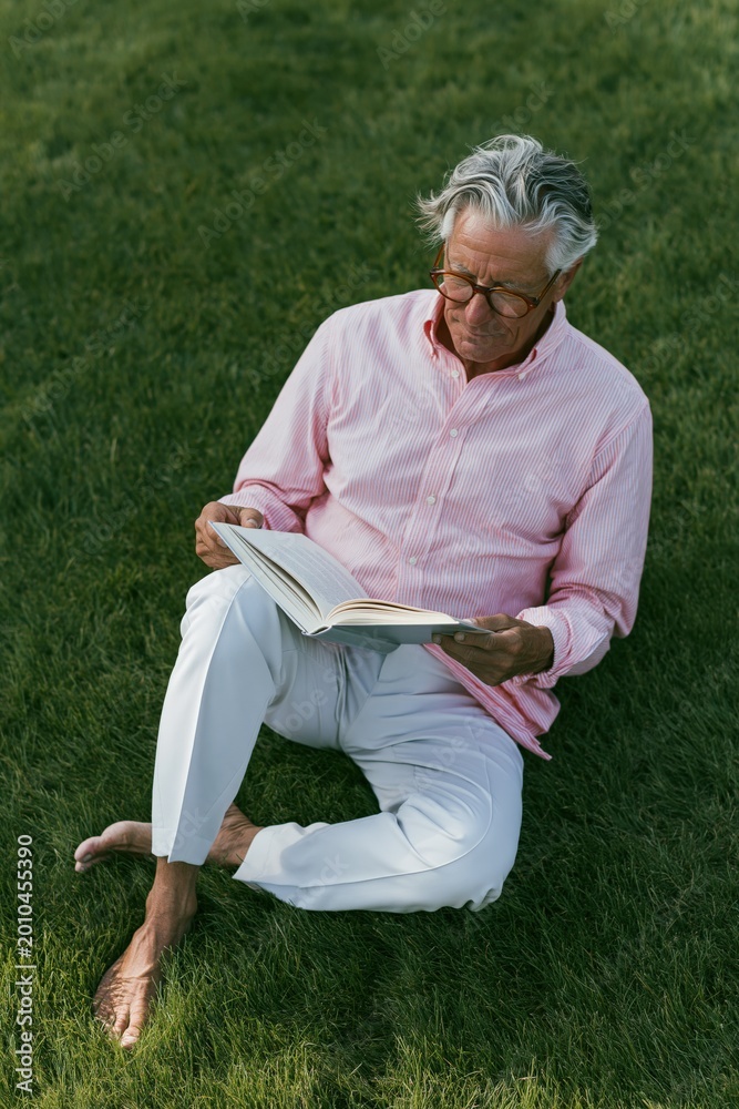 Fototapeta premium Elderly man reading open book seated on grassy ground