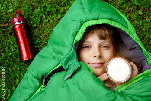 Preteen school kid boy in sleeping bag camping. Outdoors activity with children in summer. Fun and adventure camp, family and friends vacations or weekend trip. Portrait of child with flashlight.
