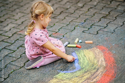 Little preschool girl painting rainbow with colorful chalks on ground on backyard. Positive happy toddler child drawing and creating pictures. Creative outdoors activity in summer.