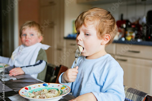 Two little boys eating meal in kitchen at home or daycare nursery, siblings enjoying food together at table, candid family lifestyle moment, real life, real people.
