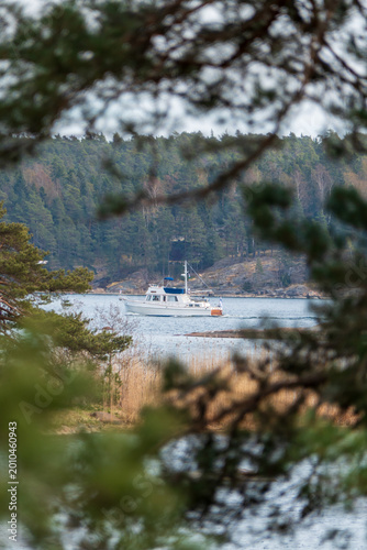 Motorboat cruising on a tranquil bay surrounded by forest, seen through soft-focus tree branches. Natural framing and muted tones evoke a calm, remote Scandinavian coastal setting.