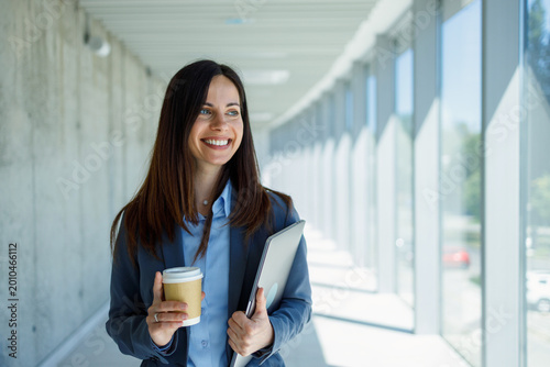 Businesswoman walking in modern office hallway holding coffee and laptop